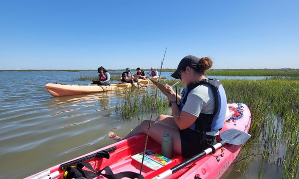 Artist Boat kayak tours in Galveston, TX. Girl sitting in kayak while drawing with another kayak in background.